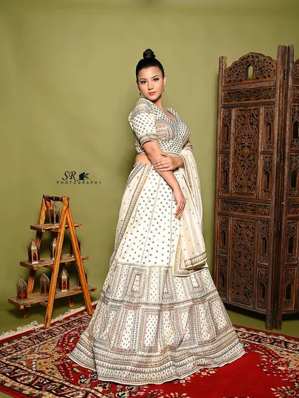 A full-length portrait of a model in an elegant white and gold lehenga. The composition uses the decorative ladder and rug to frame the shot, creating a complete, styled scene.