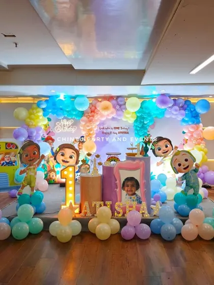 A large-scale Cocomelon themed first birthday stage decoration. The pastel rainbow balloon arch and multiple character cutouts create an immersive experience.
