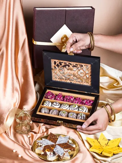 Hands opening a luxurious, dark wood gift box to reveal rows of colorful Indian sweets. This shot creates a sense of anticipation and premium gifting.