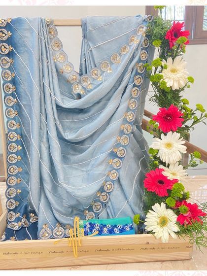A beautiful blue saree with intricate border work, elegantly displayed on a wooden trousseau stand adorned with fresh gerberas and greenery.