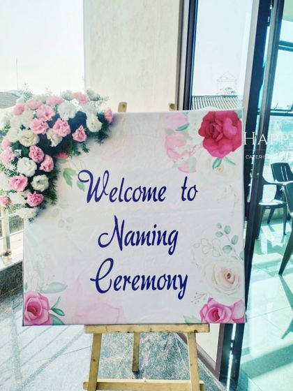 A simple and elegant welcome sign for a naming ceremony, placed on an easel. The sign is decorated with a watercolor floral design and a fresh flower accent.
