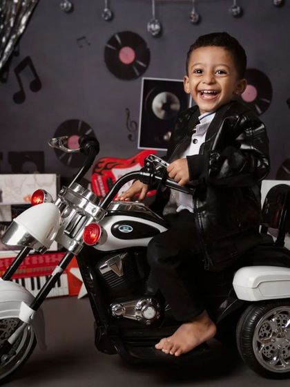 This toddler is all smiles in a rock-and-roll biker themed shoot. Dressed in a leather jacket, he sits on a toy motorcycle against a backdrop of vinyl records and silver streamers.
