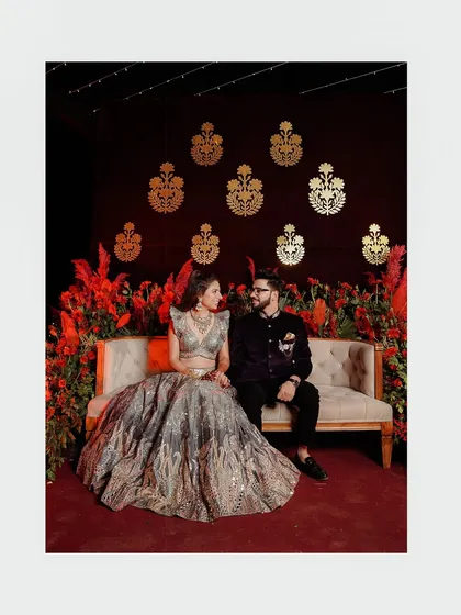 A couple sits on a couch against a backdrop of rich red flowers and golden motifs, creating a regal and romantic portrait from their Qawali night.