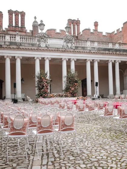 A wide shot of the ceremony setup, showing the rows of chairs facing the magnificent floral mandap. The composition highlights the grandeur of an outdoor wedding in a royal courtyard.