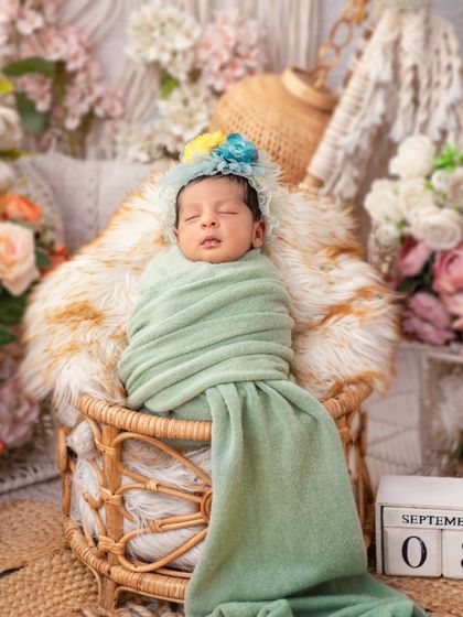 A newborn wrapped in a soft green blanket rests in a boho-style wicker chair, surrounded by pampas grass and muted floral tones. This setup is perfect for families who love a natural, earthy, and chic aesthetic.