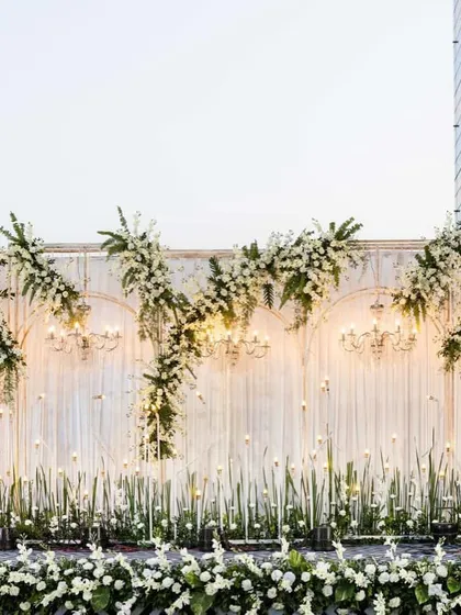 An elegant, all-white stage backdrop for a reception or Sangeet. The design uses drapery, chandeliers, and floral arrangements to create a sophisticated and classic look.