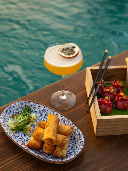 A beautiful spread of Pan-Asian food and cocktails by the water. This shot captures a complete dining experience, perfect for a high-end beach club or restaurant.