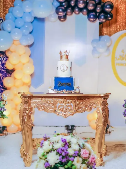 The royal cake display, featuring a magnificent two-tiered cake with a crown topper, placed on an ornate golden table. The backdrop of balloons and shimmering curtains adds to the grandeur.