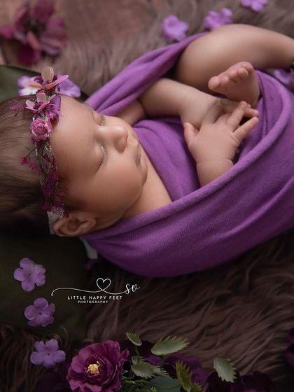 A close-up on a baby girl wrapped in purple, surrounded by matching flowers. The focus is on her peaceful expression and the delicate floral headband.