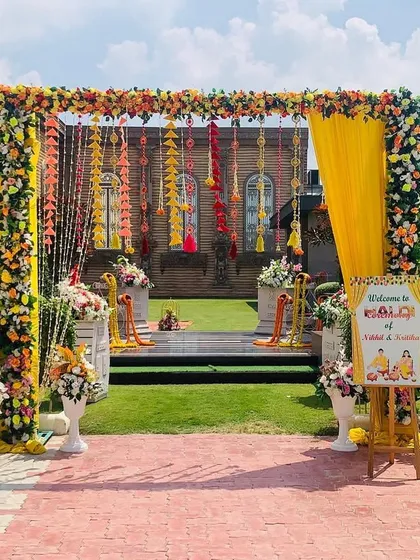 A vibrant and traditional Haldi ceremony entrance. This setup features a beautiful arch made of fresh marigolds and other colorful flowers, with hanging floral strings and a personalized welcome sign for the couple.