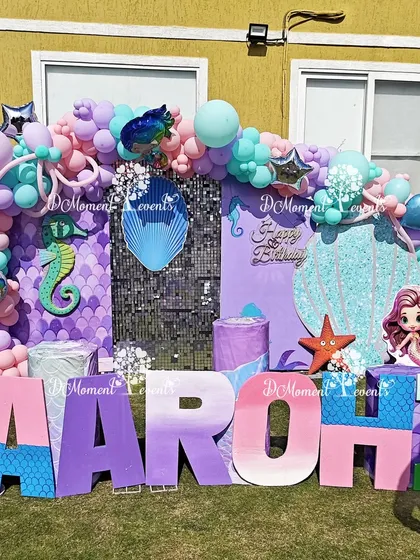A vibrant mermaid-themed first birthday stage set up on a lawn. The decor includes large marquee letters spelling "AAROHI," a sequin backdrop, and numerous under-the-sea character cutouts and props.