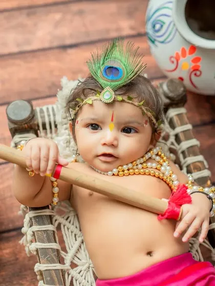 A close-up shot of a baby dressed as Krishna, lying on a miniature cot and holding a flute.