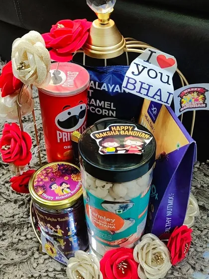 A premium Rakhi hamper in a golden birdcage-style basket, filled with Pringles, caramel popcorn, healthy nut mix, and other treats.