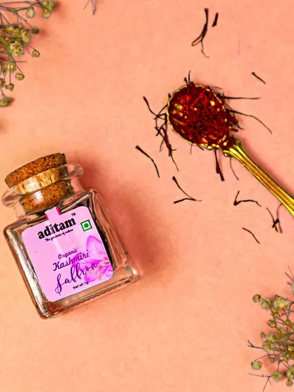 The luxuriousness of Kashmiri saffron is captured in this beautiful flatlay. The vibrant red strands are displayed in a glass jar and on an ornate golden spoon against a soft pink background.