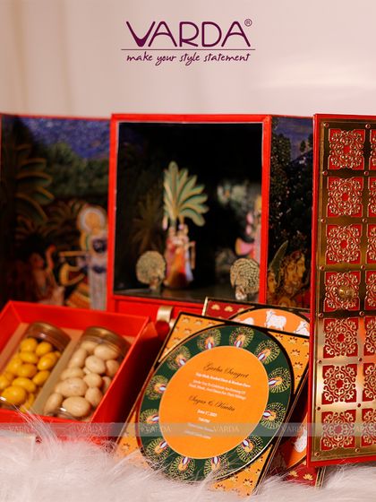 A spectacular multi-panel invitation box featuring Pichwai art. It opens up to reveal compartments for gourmet gifts and a series of beautifully designed, circular wedding cards, creating a grand and artistic unboxing experience.