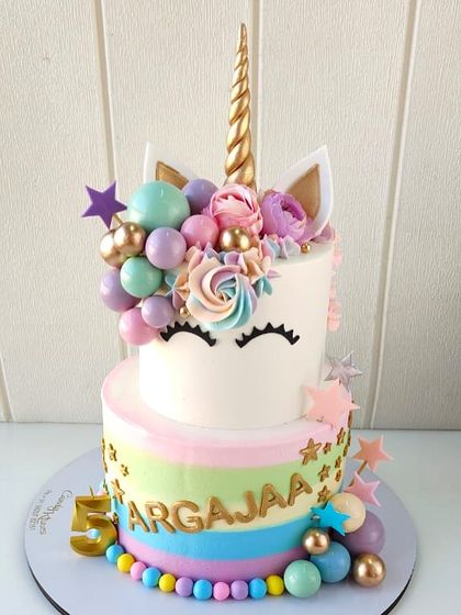 A magical unicorn cake designed for a fifth birthday celebration. This two-tiered creation features a pastel rainbow striped bottom tier, a sleepy unicorn face on the top tier, and is decorated with a golden horn, floral details, and candy-colored spheres.
