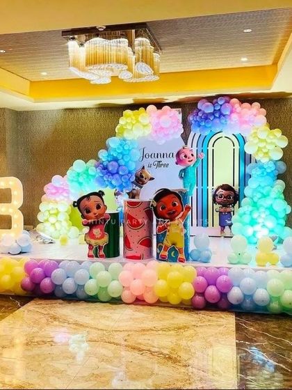 A vibrant Cocomelon-themed stage decoration for a third birthday. The setup features character cutouts, a large light-up marquee number '3', and a multi-level stage decorated with pastel rainbow balloons.
