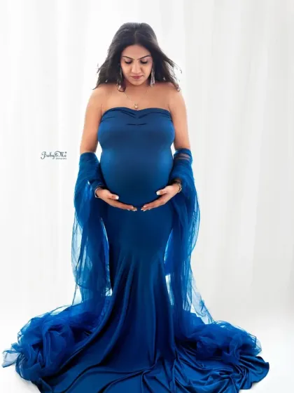 A classic and elegant pose. This mom-to-be cradles her bump in a stunning royal blue mermaid gown with a sheer wrap, set against a clean white background for a timeless feel.