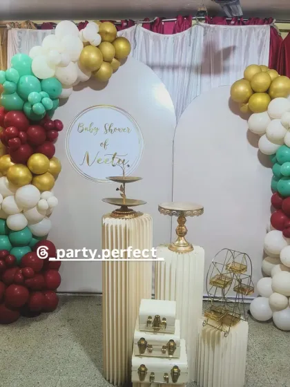 A modern baby shower backdrop with elegant white arched panels and a balloon arrangement in maroon, teal, white, and gold.