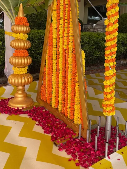 A close-up of the traditional decor elements for a Haldi ceremony, including marigold garlands, brass pots (kalash), and a vibrant yellow patterned floor.