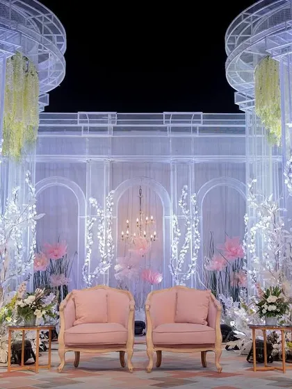 The main seating area on the reception stage, framed by ethereal white branches and soft pink chairs. The backdrop is a translucent wall that catches the light beautifully.