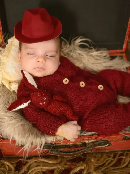 A sleeping newborn in a red knit outfit, holding a small stuffed rabbit while lying in a vintage-style suitcase.