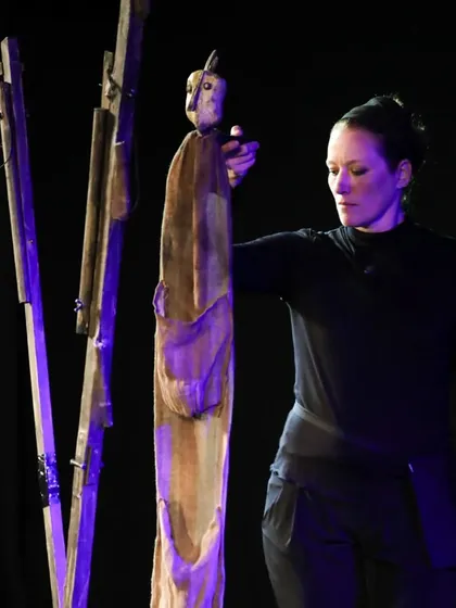 A puppeteer interacts with a tall, abstract wooden puppet in 'Wings & Roots'. This French performance uses non-traditional puppets to tell a story about peace and harmony.