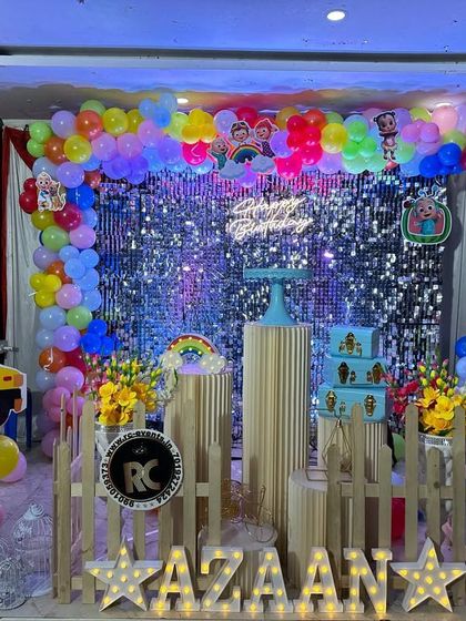 A vibrant Cocomelon setup featuring a silver shimmer wall and a colorful balloon frame. The decor includes a Cocomelon bus cutout and themed cake plinths.
