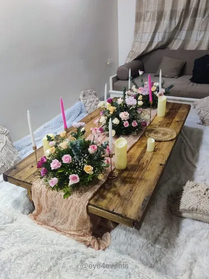 This rustic-chic picnic setup features a low wooden table on a plush white rug. The decor is enhanced with a delicate table runner, fresh floral arrangements, and macrame cushions for a cozy, bohemian feel.