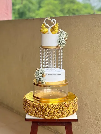 An elegant three-tier engagement cake featuring golden swans, baby's breath, and crystal separators filled with fairy lights. The base is adorned with beautiful golden roses, creating a luxurious look.