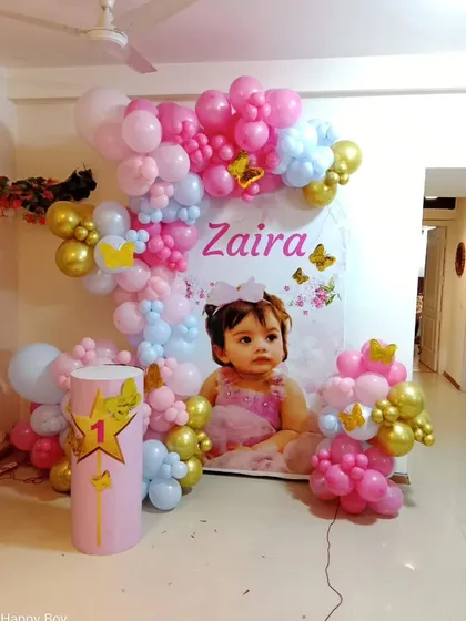 A personalized first birthday setup with a large photo of the birthday girl as the centerpiece, framed by pink, blue, and gold balloons.