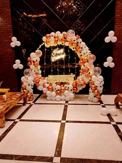A beautiful balloon arch in white and rose gold. This setup includes a neon sign and fairy lights, creating an elegant photo corner in a venue with marble floors and dark walls.