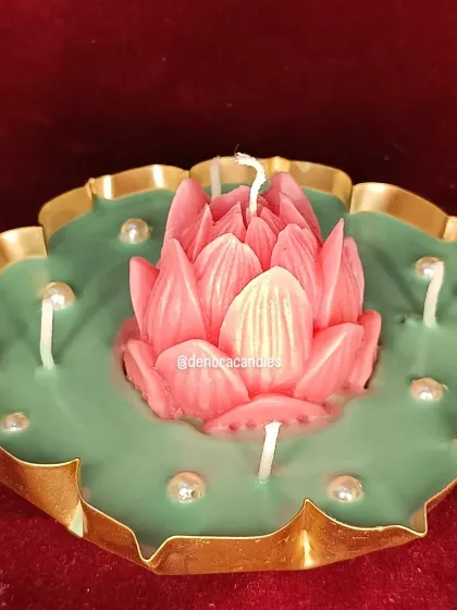This is my classic lotus flower urli candle. It features a central pink lotus sculpture on a green wax base, with multiple wicks and pearl accents, all set in a traditional brass urli.