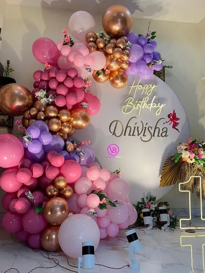 A stunning and modern birthday backdrop for a first birthday celebration. This design features a massive, asymmetrical organic balloon garland in shades of pink, purple, and rose gold, set against a clean white circular backdrop with custom gold lettering and a lit-up marquee number.
