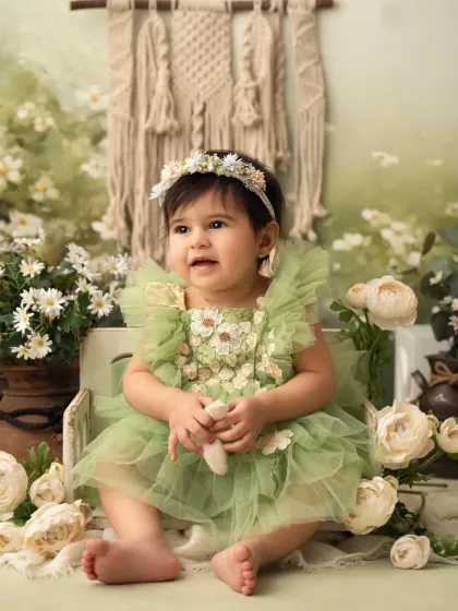 A sweet baby girl in a green dress sits in a rustic box surrounded by white flowers. The bohemian backdrop adds a lovely touch.
