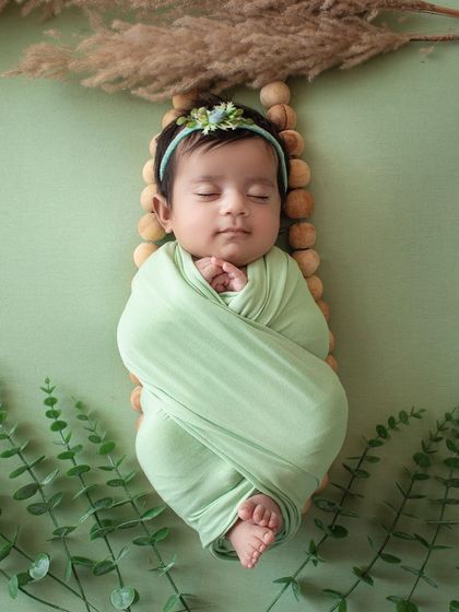 A sleeping newborn wrapped in a sage green swaddle, resting on a wooden prop with eucalyptus leaves. A very natural and calming setup.
