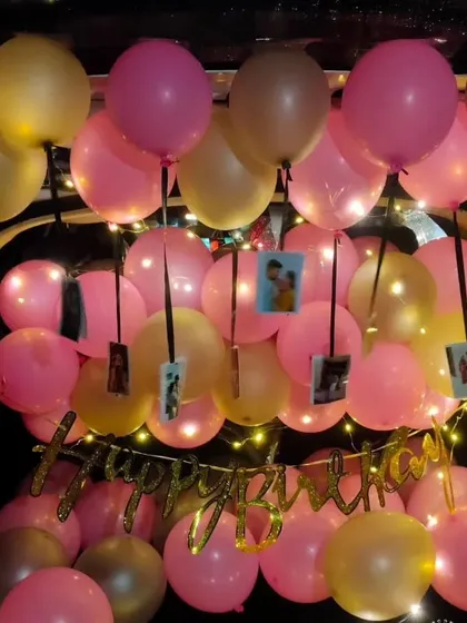 A warm and personal birthday car trunk setup. The space is filled with pink and gold balloons, with small photos hanging from ribbons among them, all illuminated by a "Happy Birthday" banner and soft fairy lights.