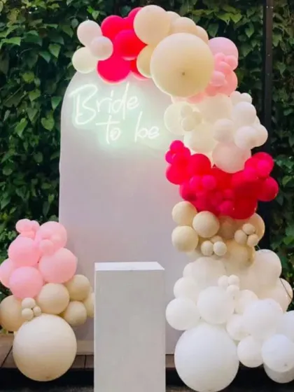 A "Bride to be" setup against a lush green foliage wall. The simple white arch with a neon sign is decorated with a modern arrangement of white and hot pink balloons.