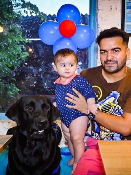 A father holds his baby next to their calm and gentle black Labrador. It's a beautiful picture of a family's love.