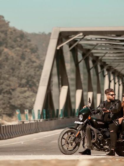 A wide shot of the couple and their motorcycle on a bridge, showcasing the beautiful mountain landscape in the background. This is a perfect example of our destination pre-wedding photography.
