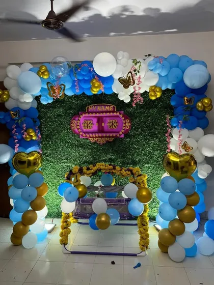 A traditional cradle ceremony decoration for a home. The setup includes a green foliage wall, a decorated cradle, and a blue, white, and gold balloon arch, blending modern and traditional elements.