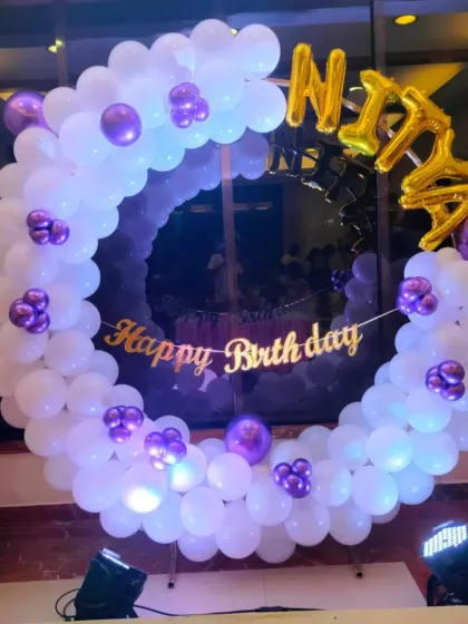 A beautiful birthday ring decoration in white and purple, perfect for a home setup. The foil name balloon and 'Happy Birthday' banner make it a personalized photo corner.