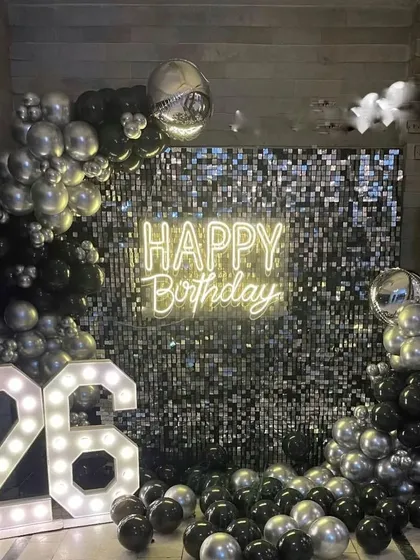 A glamorous 26th birthday party decor with a black and silver theme. The setup includes a full silver shimmer wall, a 'Happy Birthday' neon sign, large marquee numbers, and a mix of chrome and black balloons.