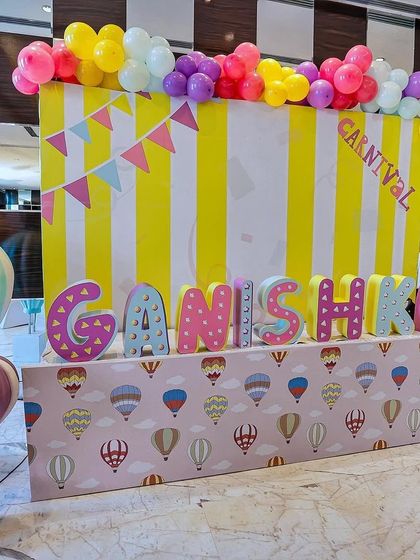 A vibrant carnival backdrop with a personalized name display for "Ganishka." The colorful letters and hot air balloon patterned table create a fun and festive atmosphere.