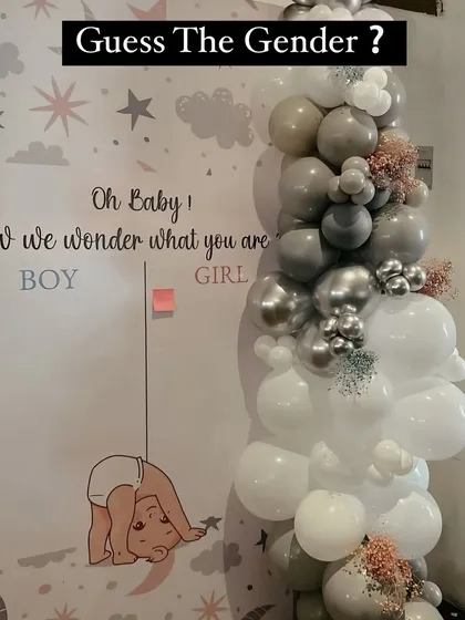 A "Guess The Gender" voting board is a fun activity for a baby shower. This one features a cute design and is placed next to a stylish grey and white balloon garland.
