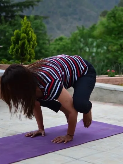 A student holds Bakasana (Crow Pose), demonstrating focus and arm strength. The supportive group energy encourages everyone to challenge themselves safely.
