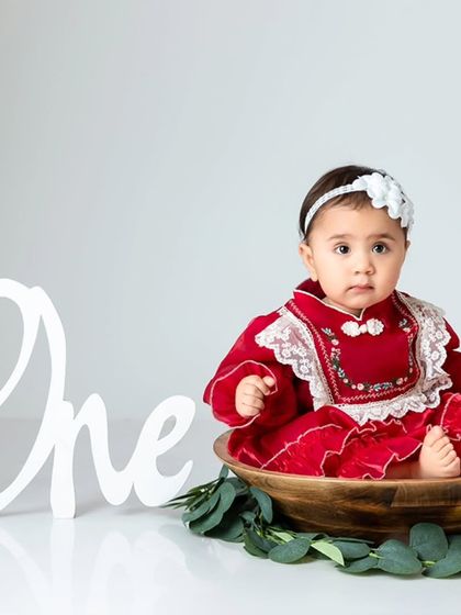 A baby girl in a red dress sits in a wooden bowl next to a "One" sign, marking her first birthday milestone.