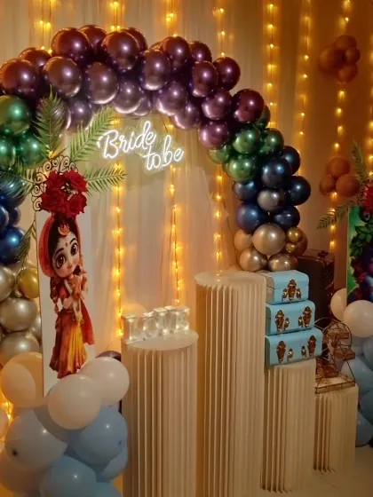 A unique bride-to-be setup with a rainbow of metallic balloons and character cutouts of Radha and Krishna. The "Bride to be" neon sign adds a modern touch to the traditional theme.