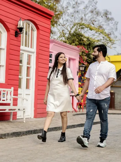 A candid walking shot from a pre-wedding session at a location with vibrant, colorful architecture. The couple is holding hands and sharing a look, creating a sense of movement and happiness as they walk toward their future together.