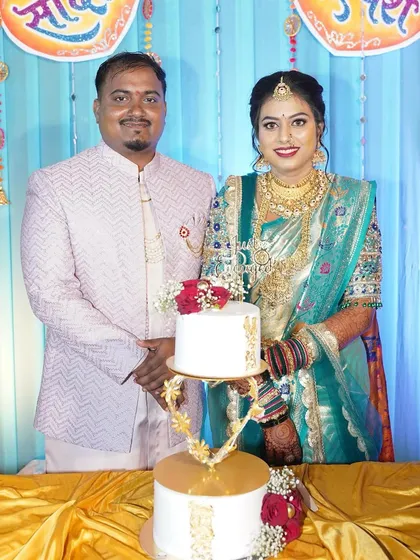 A beautiful couple stands proudly with their two-tier engagement cake, which features a unique golden stand and is decorated with fresh red roses and gold leaf accents.
